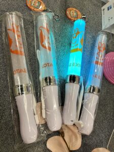 Tuotekuav Kotae Expon ensimmäisistä valotikuista. Kuvassa on 4 valotikkua kirkaissa muovisissa suojapusseissa. Valotikuissa on valkoiset kahvat, chrominen koriste kahvan ja valon liitoskohdassa. Valo-osa on koristettu Kotae Expon oranssilla logolla ja sen alla logon teksti osa. 1 neljästä valotikussa palaa turkoosivalo.